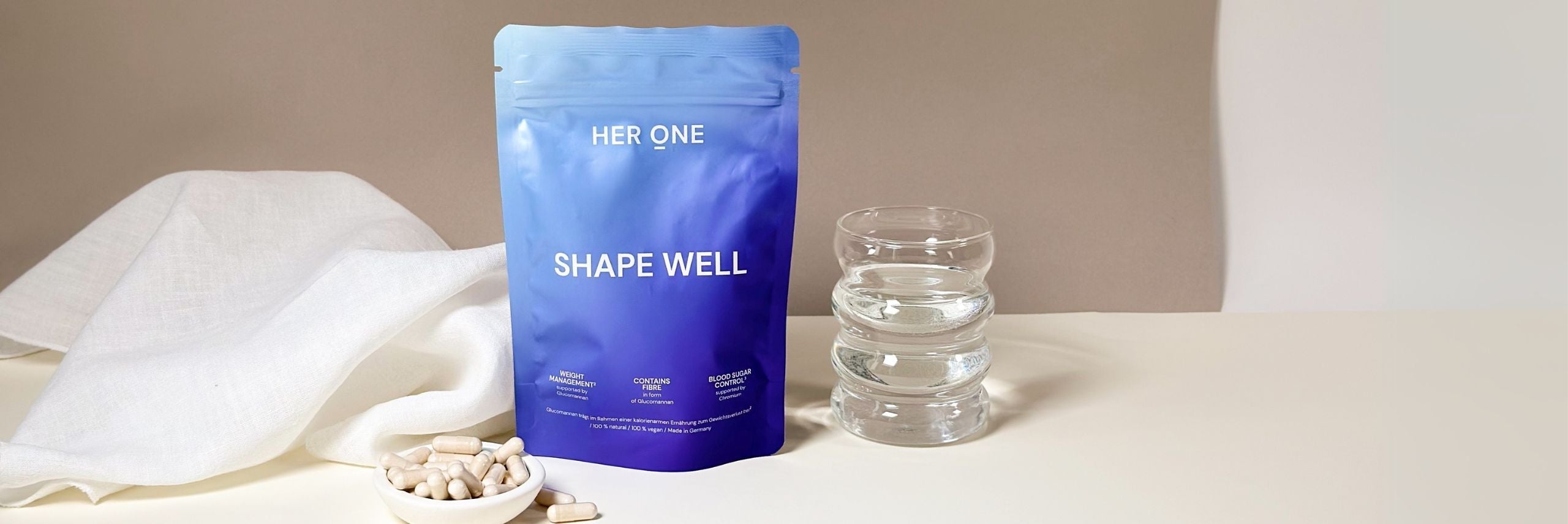 Blauer Beutel mit der Aufschrift "SHAPE WELL" neben einem Glas Wasser, einer Schale Kapseln und einem weißen Tuch.