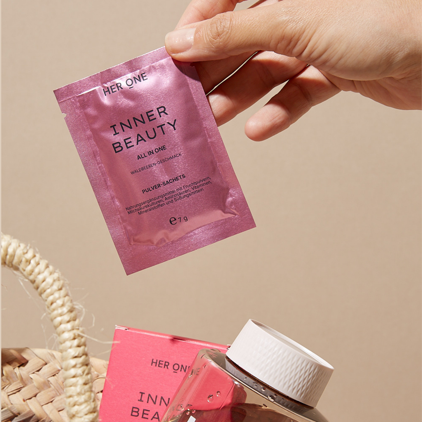 Hand hält ein rosafarbenes Sachet von Her One Inner Beauty All-in-One mit Waldbeerengeschmack. Im Vordergrund ein Shaker und eine Produktverpackung in einer Tasche.