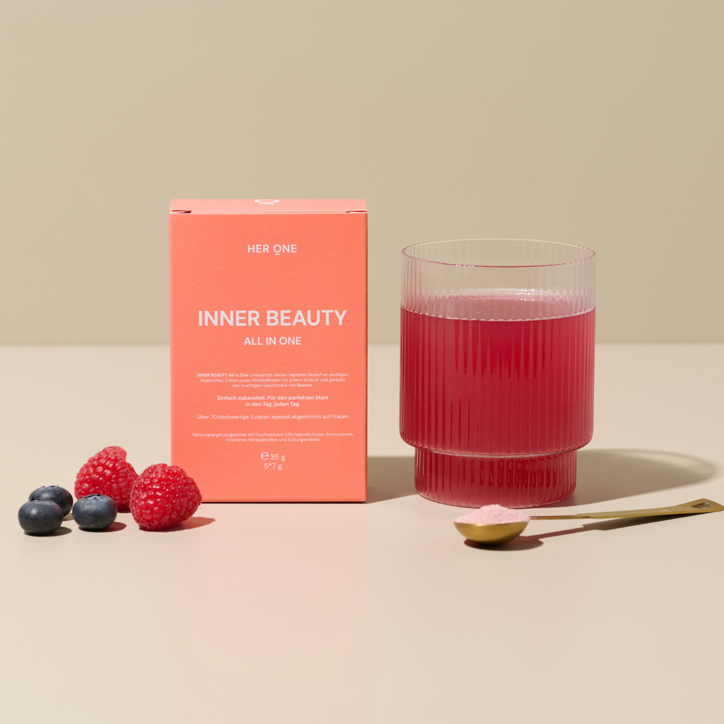 Ein Glas mit rotem Getränk, eine Packung "Inner Beauty", frische Beeren und ein Löffel mit Pulver auf einem Tisch.
