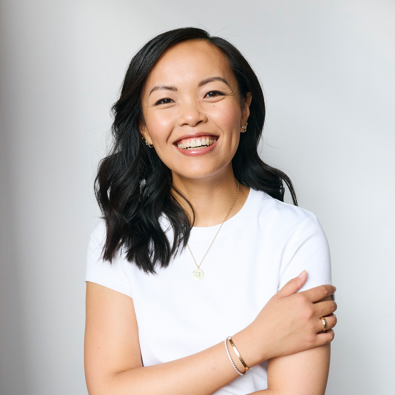 Femme souriante aux cheveux foncés portant un t-shirt blanc et des bijoux, posant devant un fond clair.