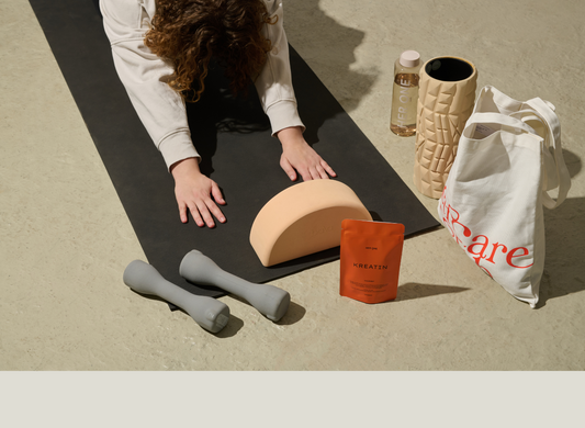 Person on yoga mat with dumbbells, bag, water bottle, half-circle block and creatine powder.
