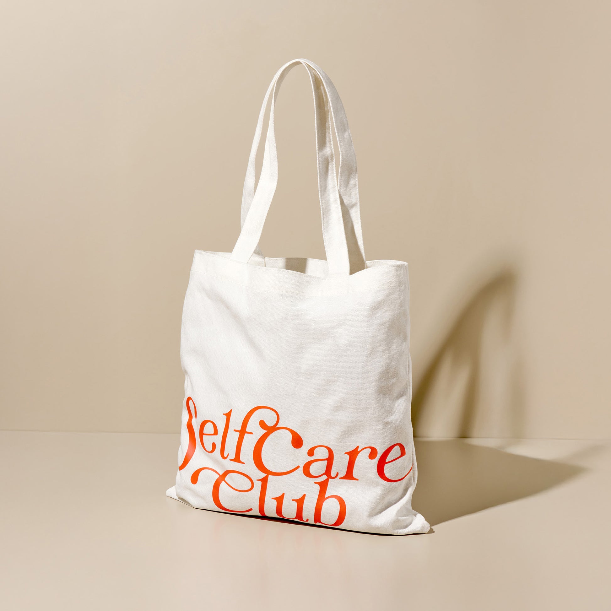 White fabric bag with orange "Self Care Club" lettering on a neutral background.