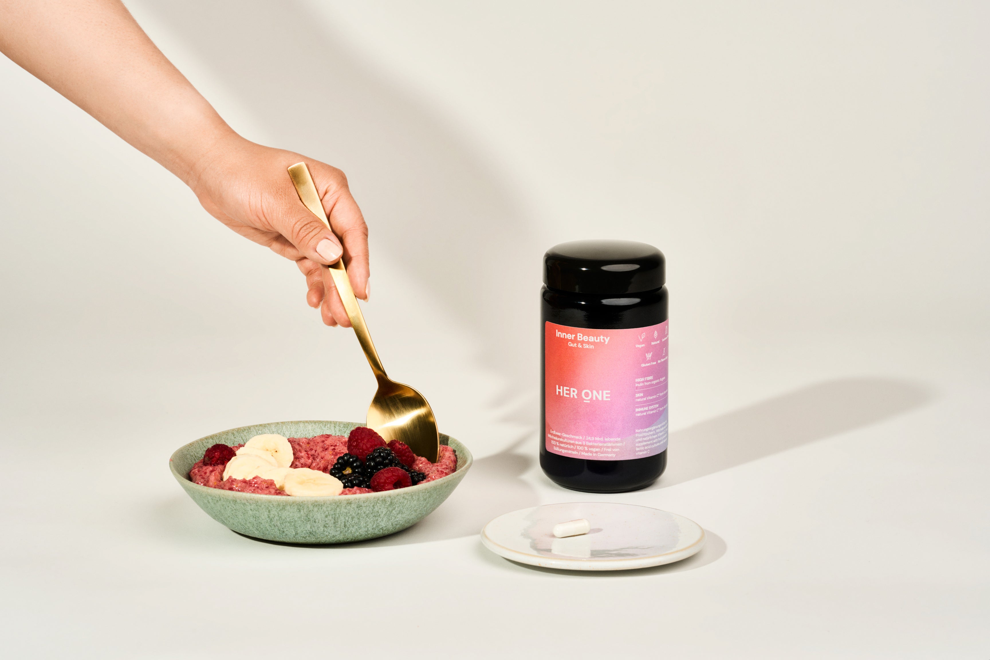 A hand with a golden spoon grabs a bowl of berries and bananas next to a food supplement.