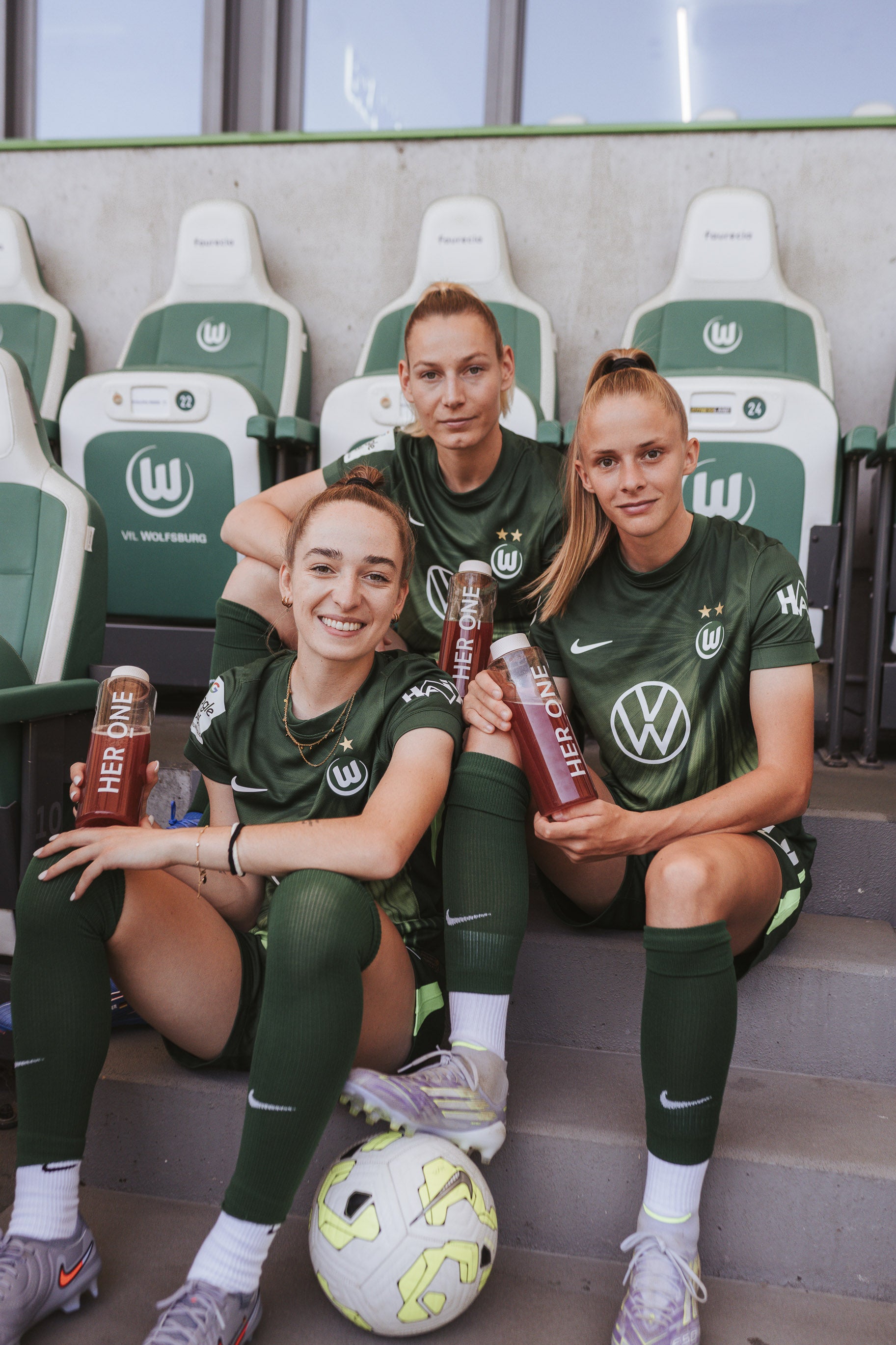 Three VfL Wolfsburg players sit on the bench and show off theirHERONEdrinkbottle.