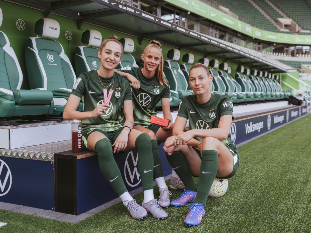 Three VfL Wolfsburg players sit on the sidelines holding HERONE products in their hands.