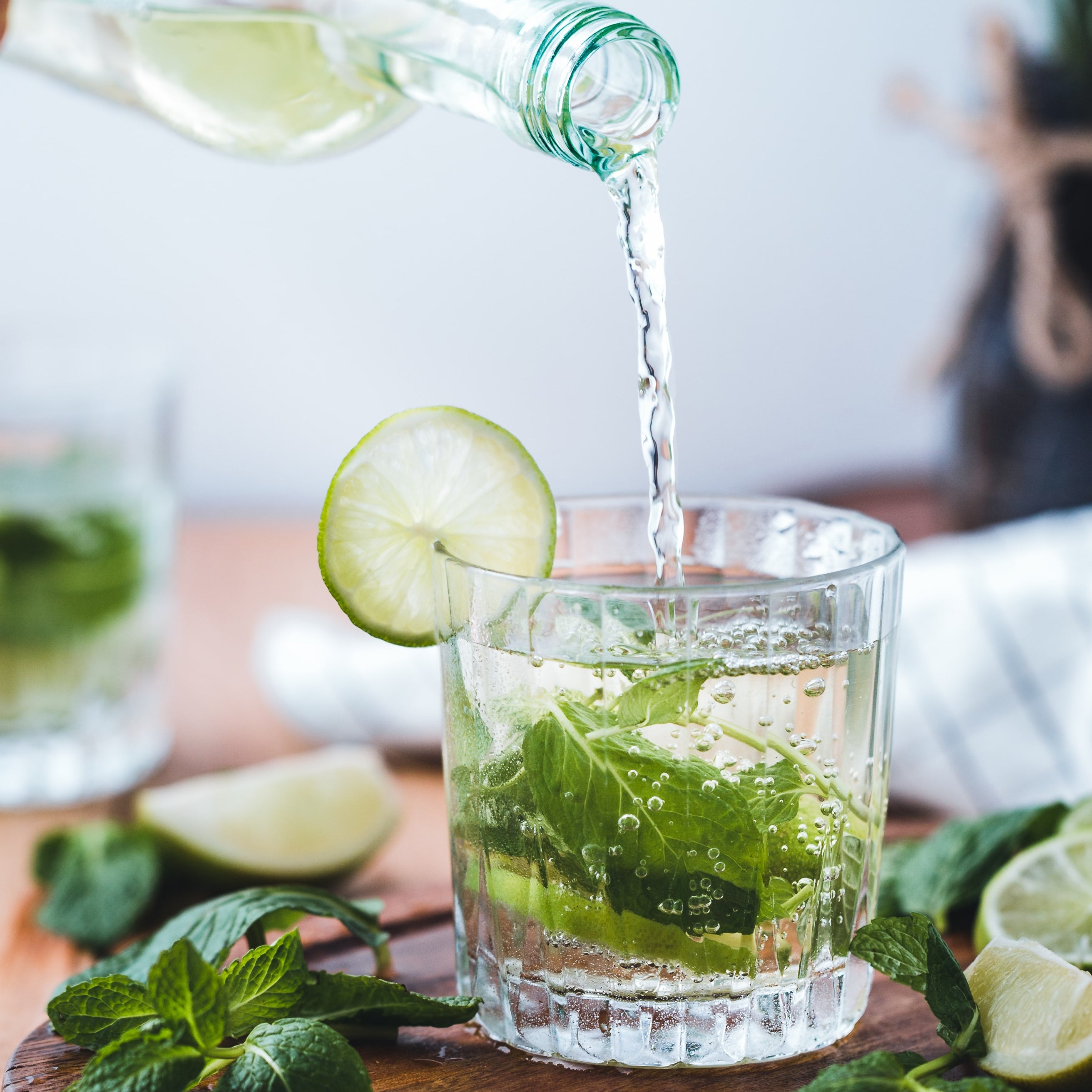 Water is poured into a glass with a slice of lime and mint, surrounded by fresh herbs on a wooden table.