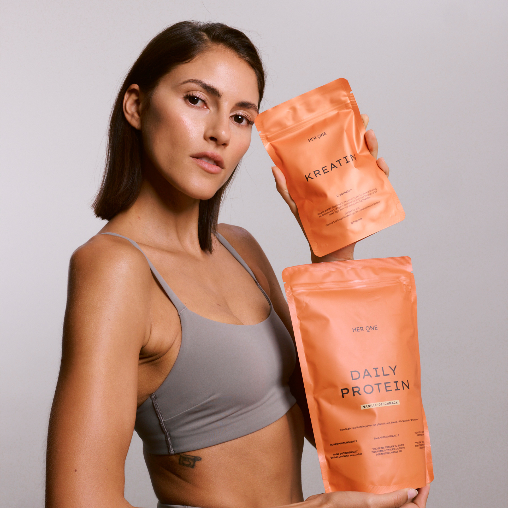 Une femme en débardeur de sport tient dans ses mains deux sachets orange HER ONE portant l'inscription Daily Protein et Kreatin.
