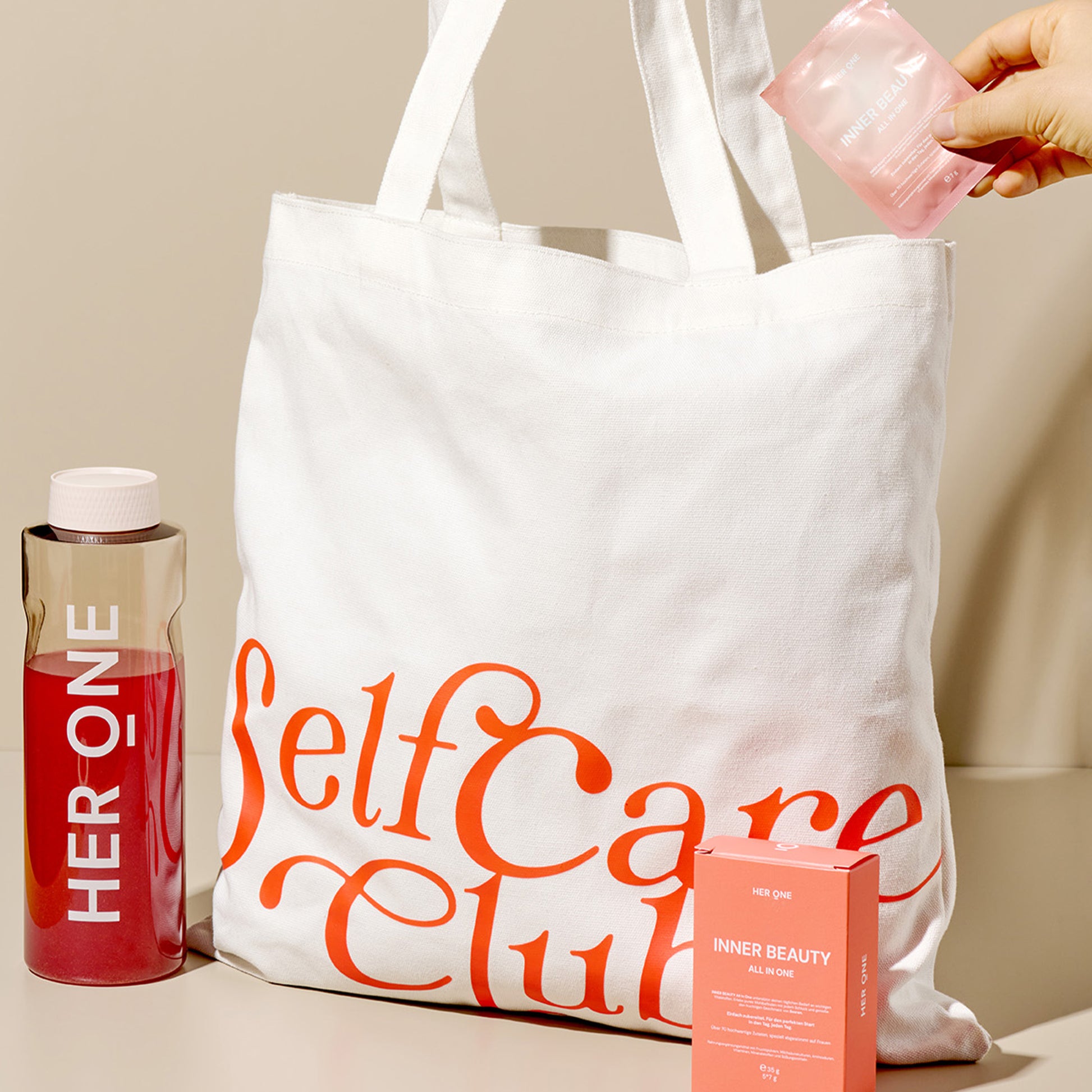 Borsa di stoffa bianca con l'etichetta "Self Care Club", accanto una bottiglia e una scatola, una mano che tiene una bustina.