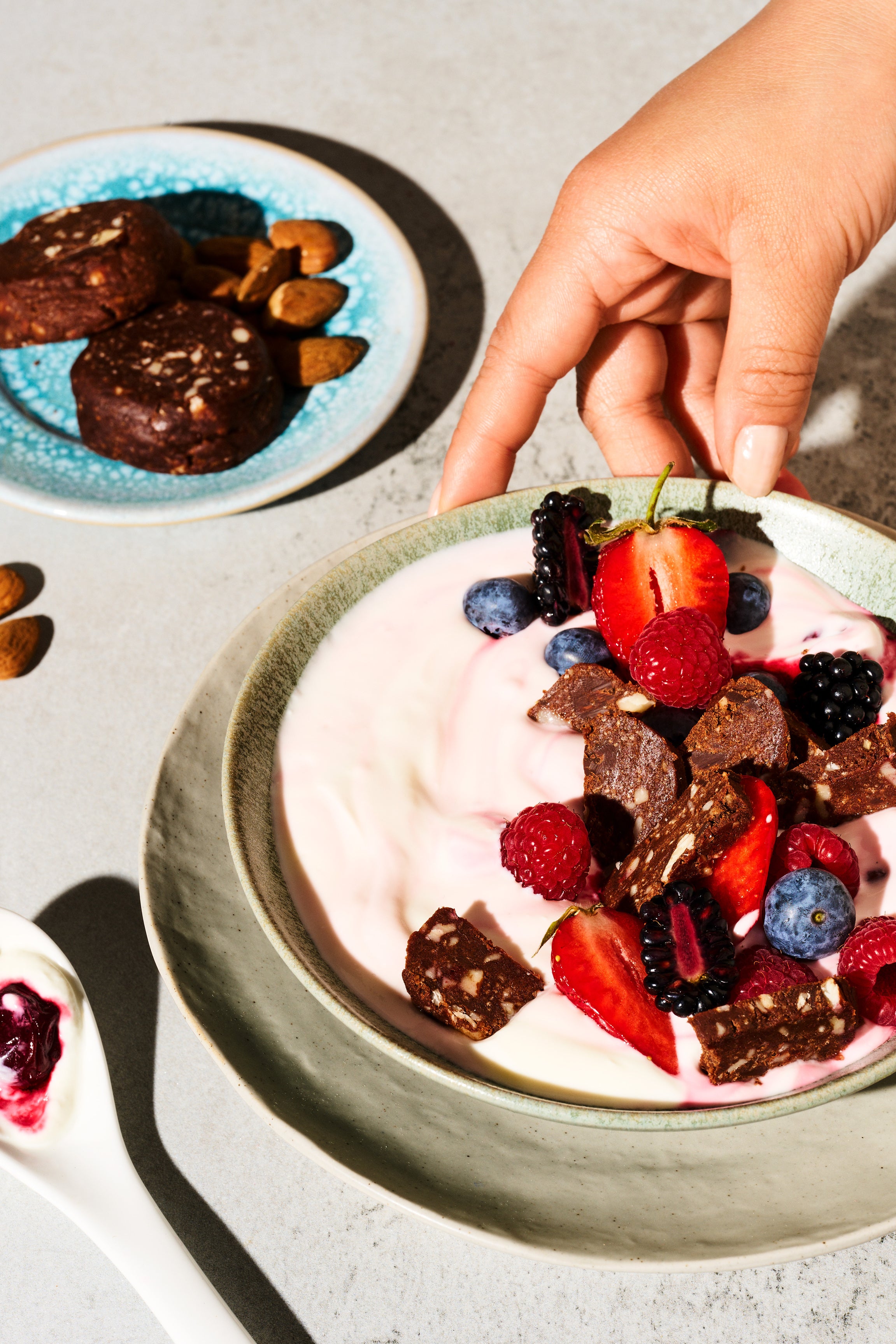 Una mano afferra una ciotola di yogurt, frutti di bosco e gocce di cioccolato; accanto un piatto di biscotti e mandorle.