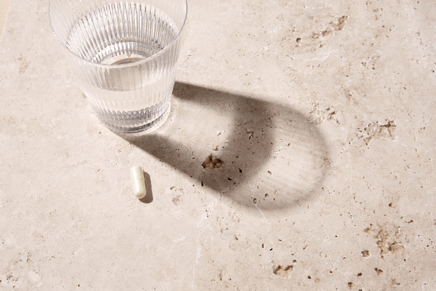 Een glas water en een capsule liggen op een lichtgekleurd stenen oppervlak.