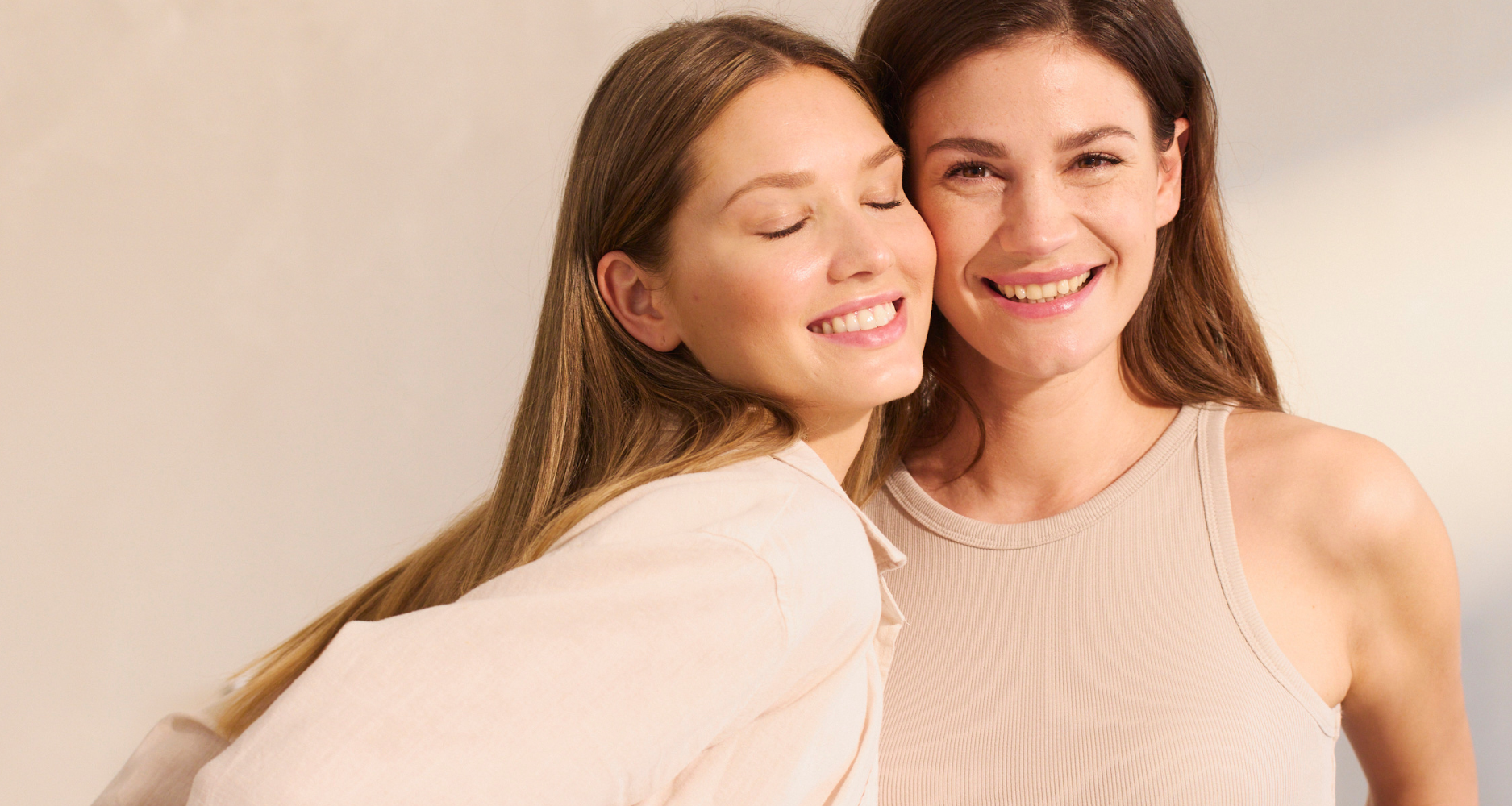 Twee lachende vrouwen in beige topjes poseren dicht bij elkaar tegen een heldere achtergrond.