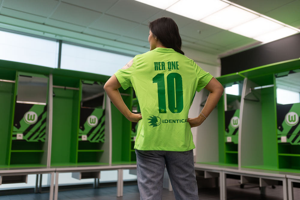Una donna con una maglia da calcio verde con il numero 10 si trova in uno spogliatoio e guarda gli armadietti aperti.