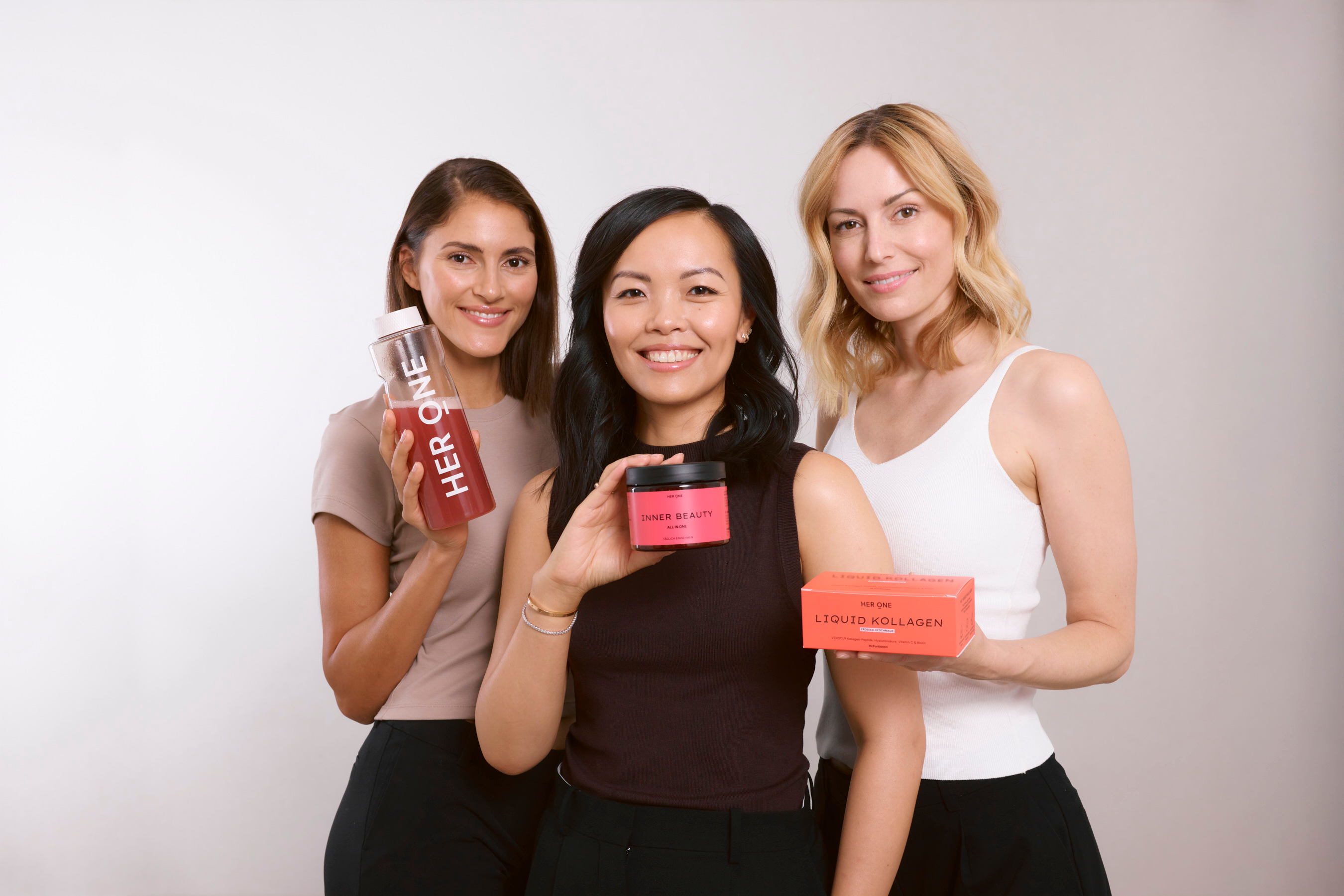 Drei Frauen präsentieren die HER ONE Produktwelt, bestehend aus Liquid Kollagen, Inner Beauty und der passenden Trinkflasche.
