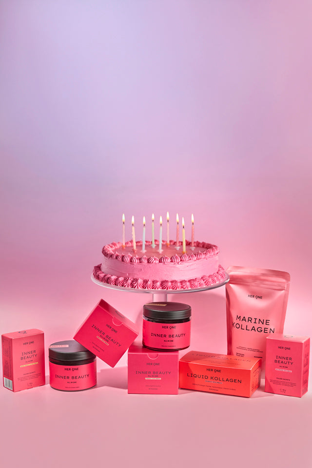 HER ONE Produkte vor einer Geburtstagstorte auf rosa-violettem Hintergrund.