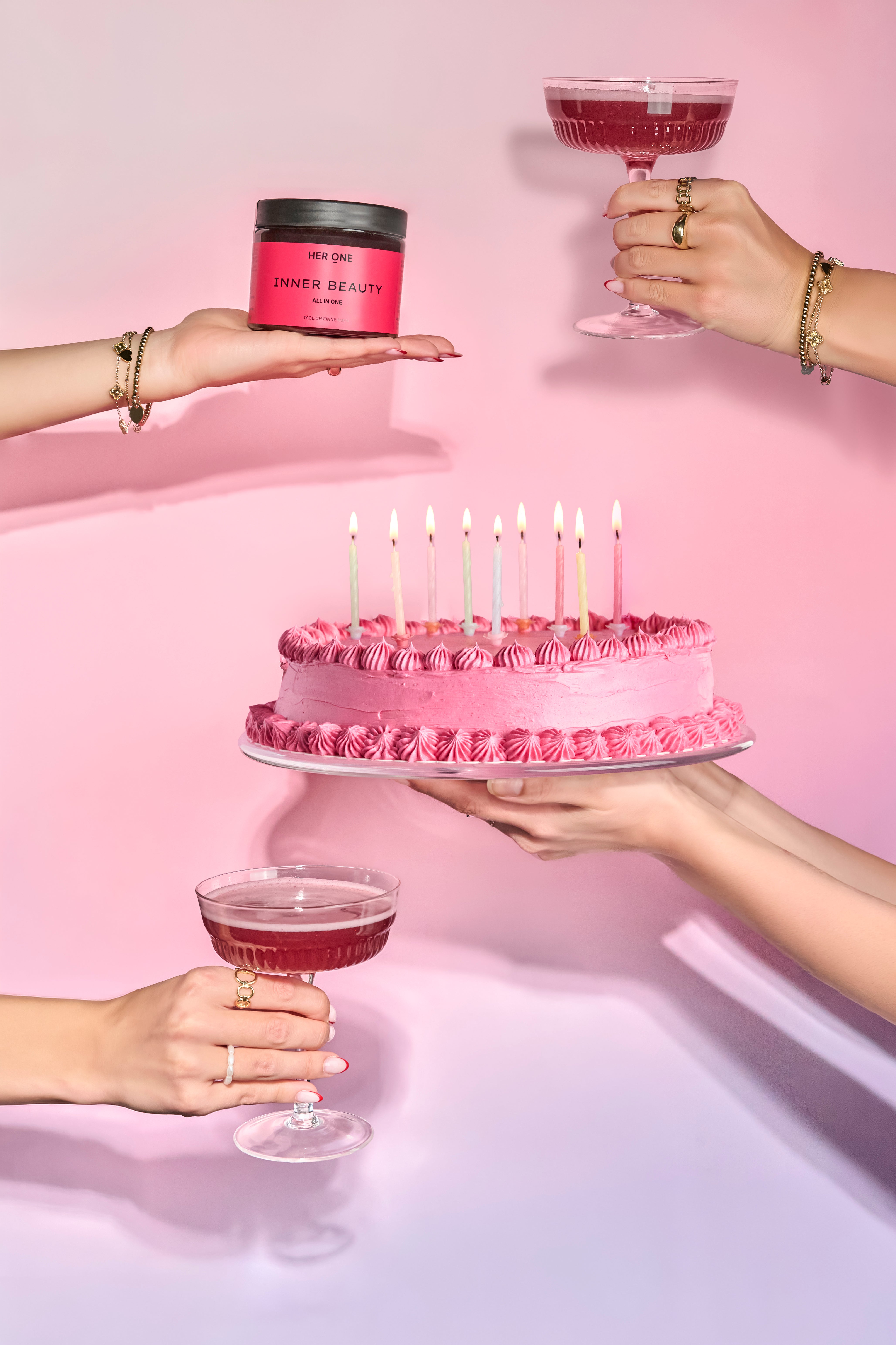 Des mains tenant un gâteau rose orné de 9 bougies, des cocktails et une boîte de HER ONE INNER BEAUTY un fond rose clair.
