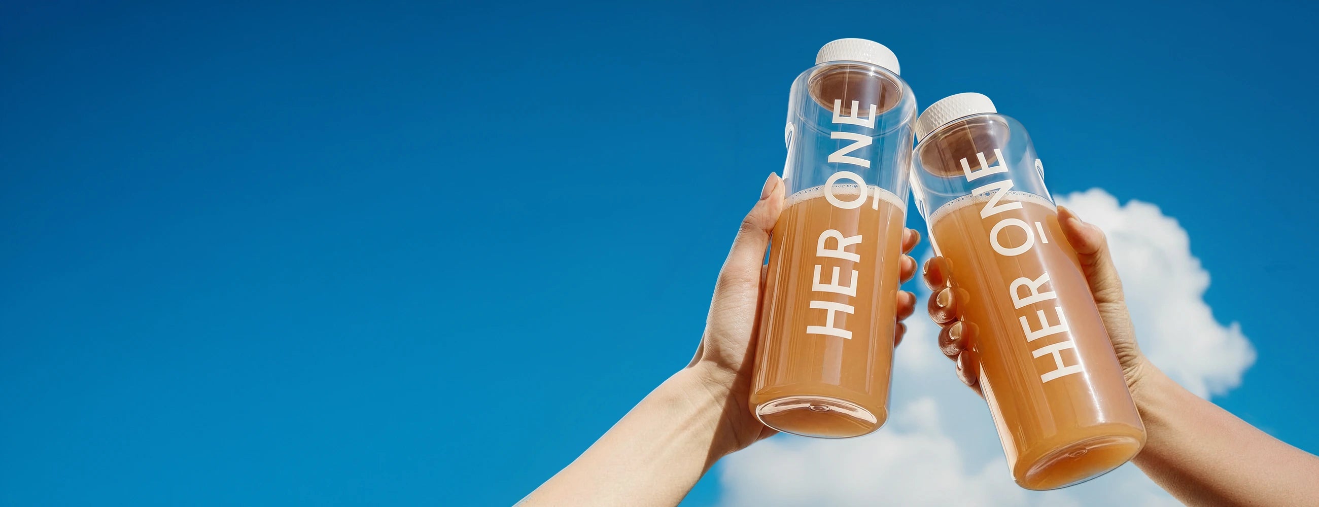 Two hands raise clear "HER ONE" bottles of INNER BEAUTY in a toast against a blue sky.