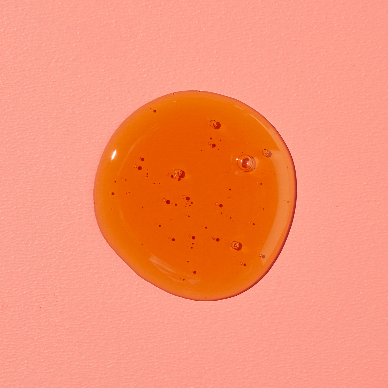 Nahaufnahme eines honigfarbenen Gel-Tropfens mit kleinen Luftbläschen auf korallrosa Fläche.