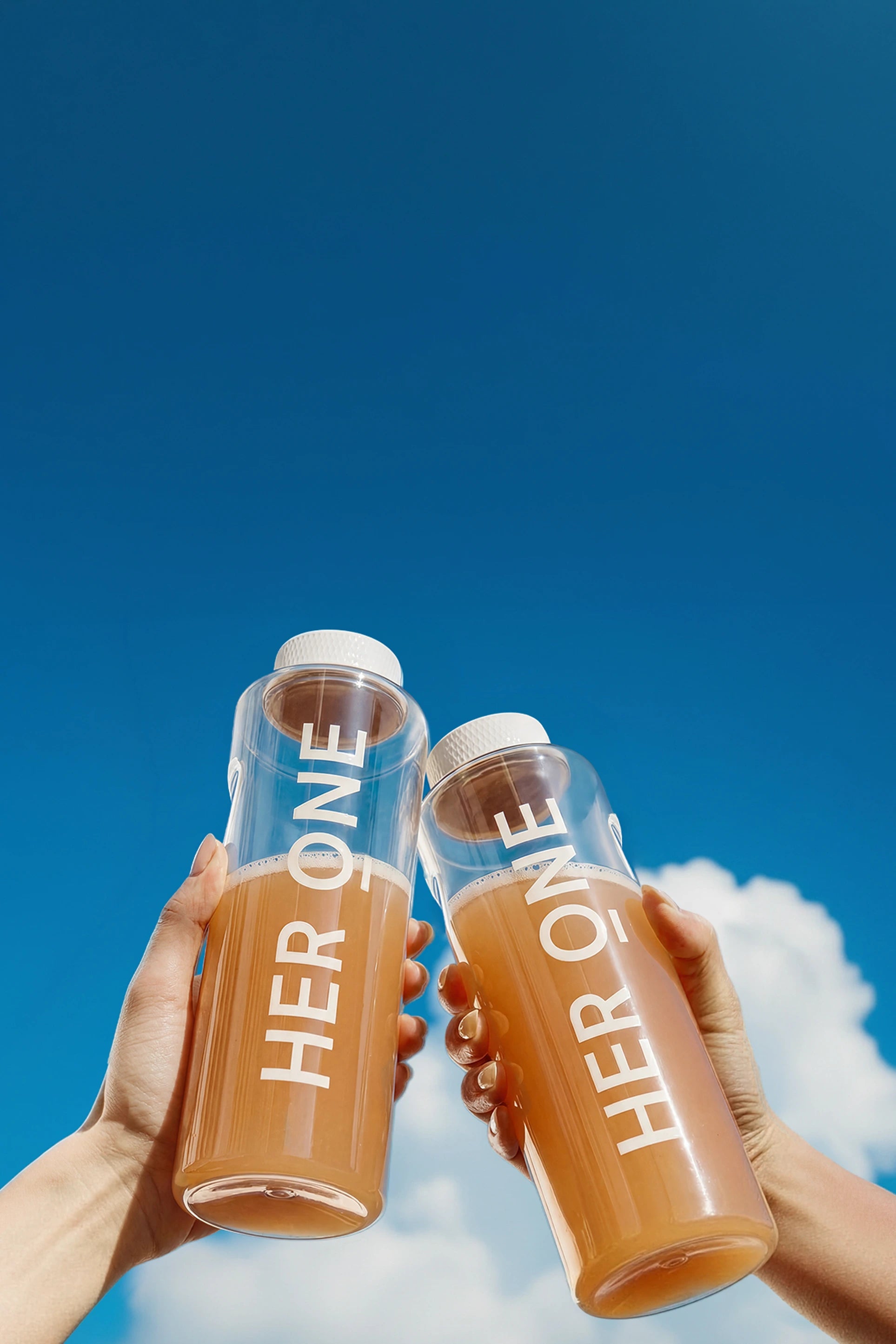 Two hands raise clear "HER ONE" bottles of INNER BEAUTY in a toast against a blue sky.