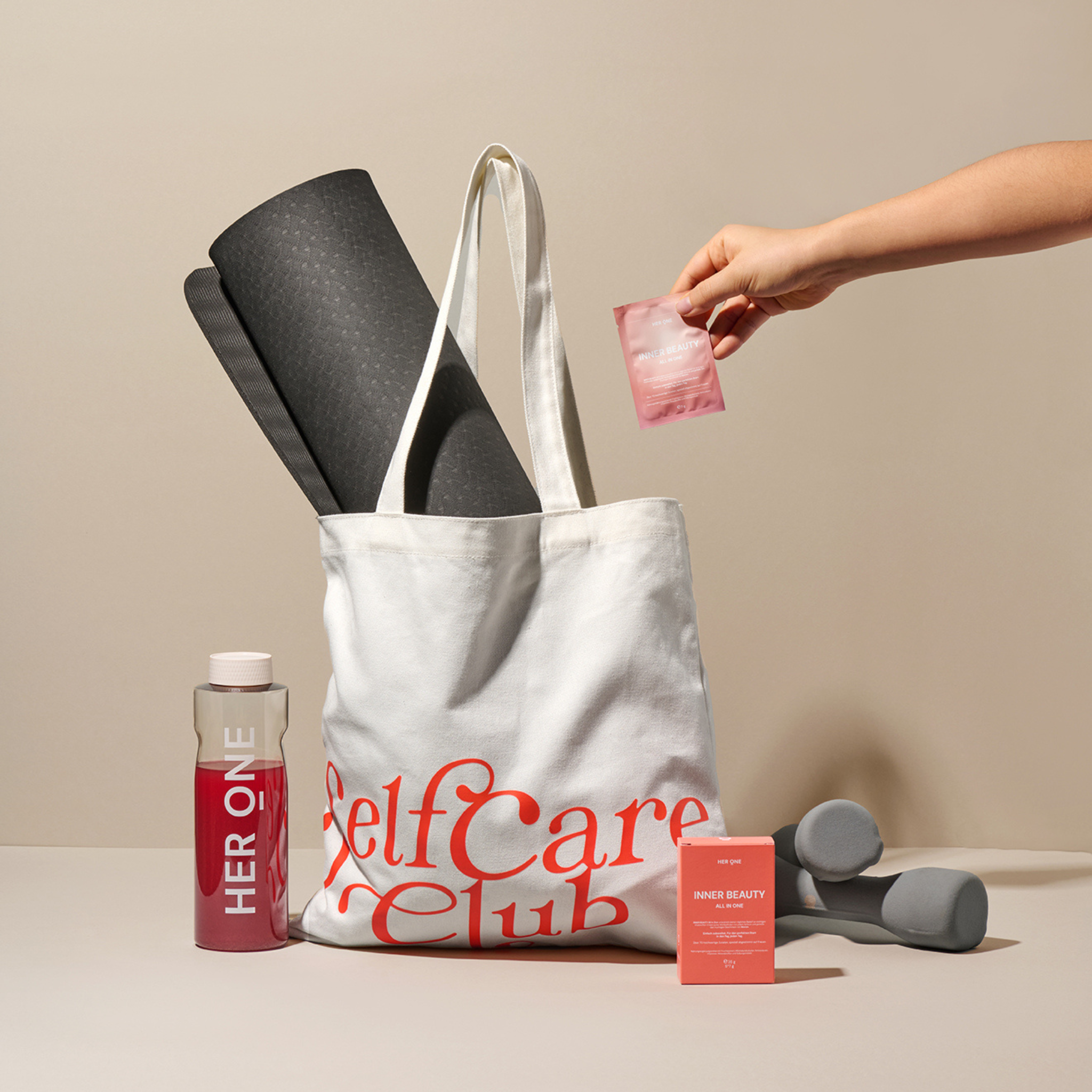 Eine Stofftasche mit "Self Care Club" bedruckt, enthält eine Yogamatte; daneben stehen Hanteln und Produkte.