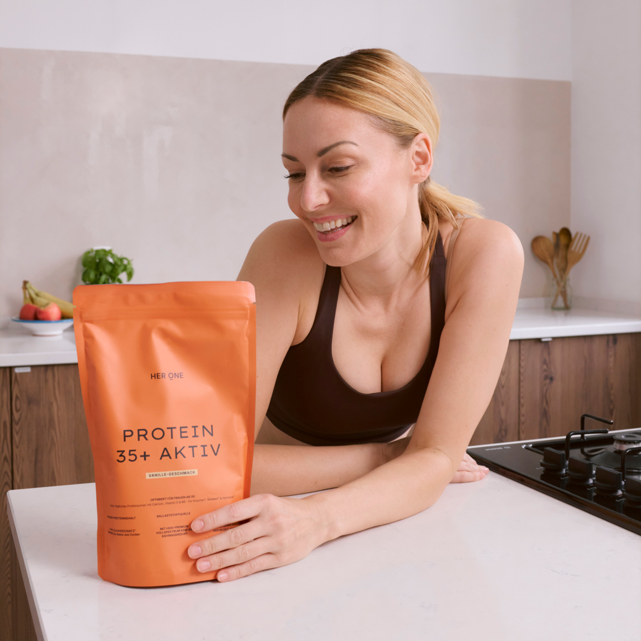 FrauindunklemSporttoplehntsichlächelndüberKüchenthekeundbetrachtetHERONEProtein35+AktivBeutel.