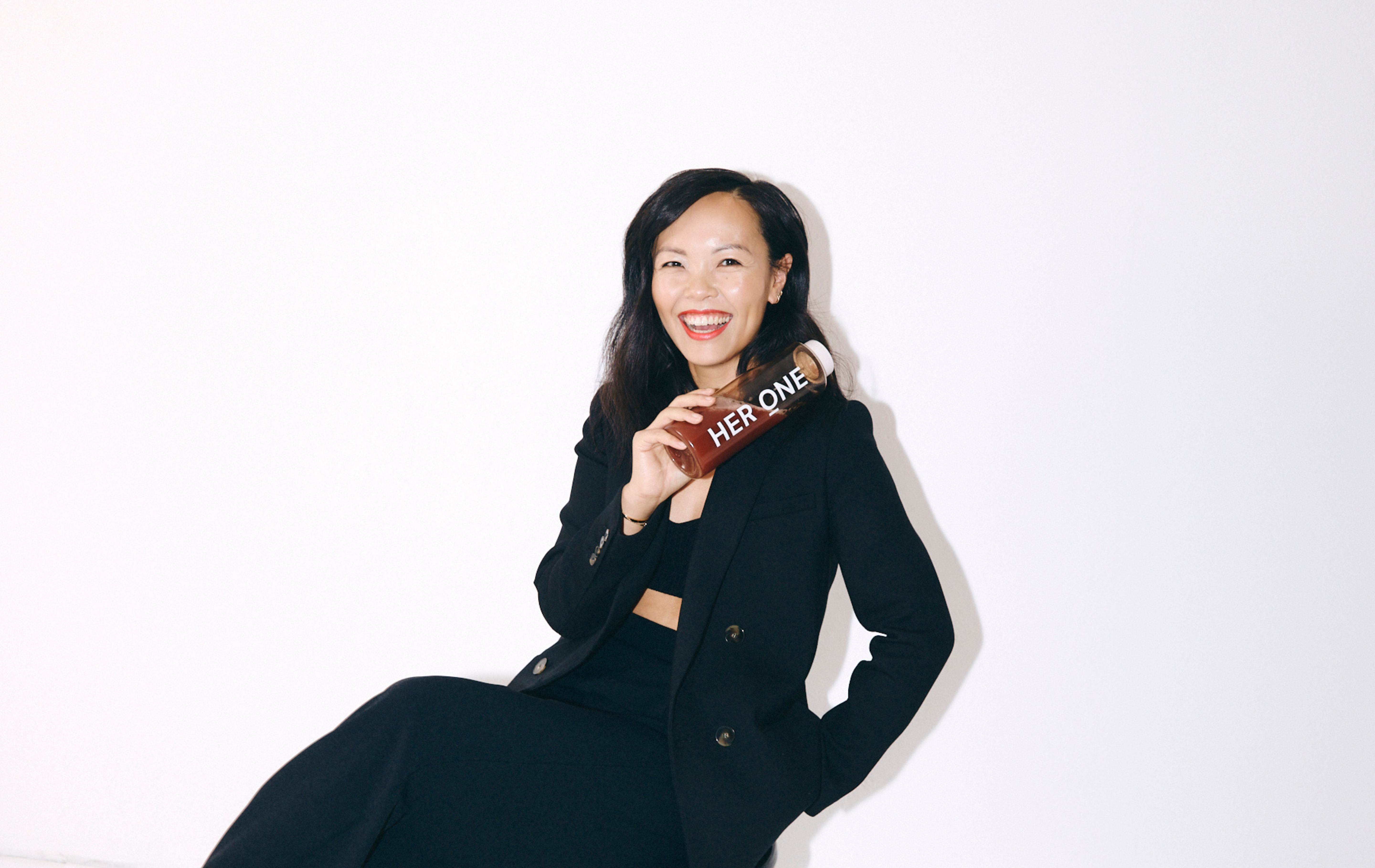 Frau in schwarzem Anzug lächelt und hält eine Flasche mit der Aufschrift "HER ONE" vor einem weißen Hintergrund.
