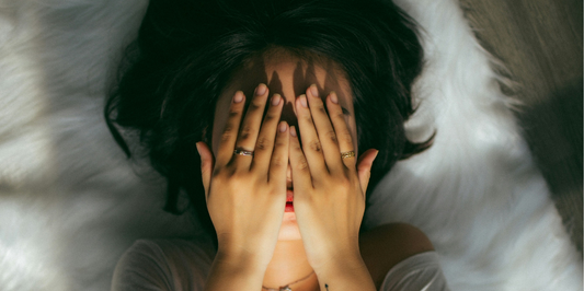 Person lies on a white fur rug and covers their face with both hands.