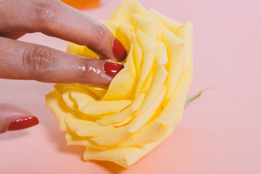 A hand with red fingernails touches a yellow rose on a pink background.