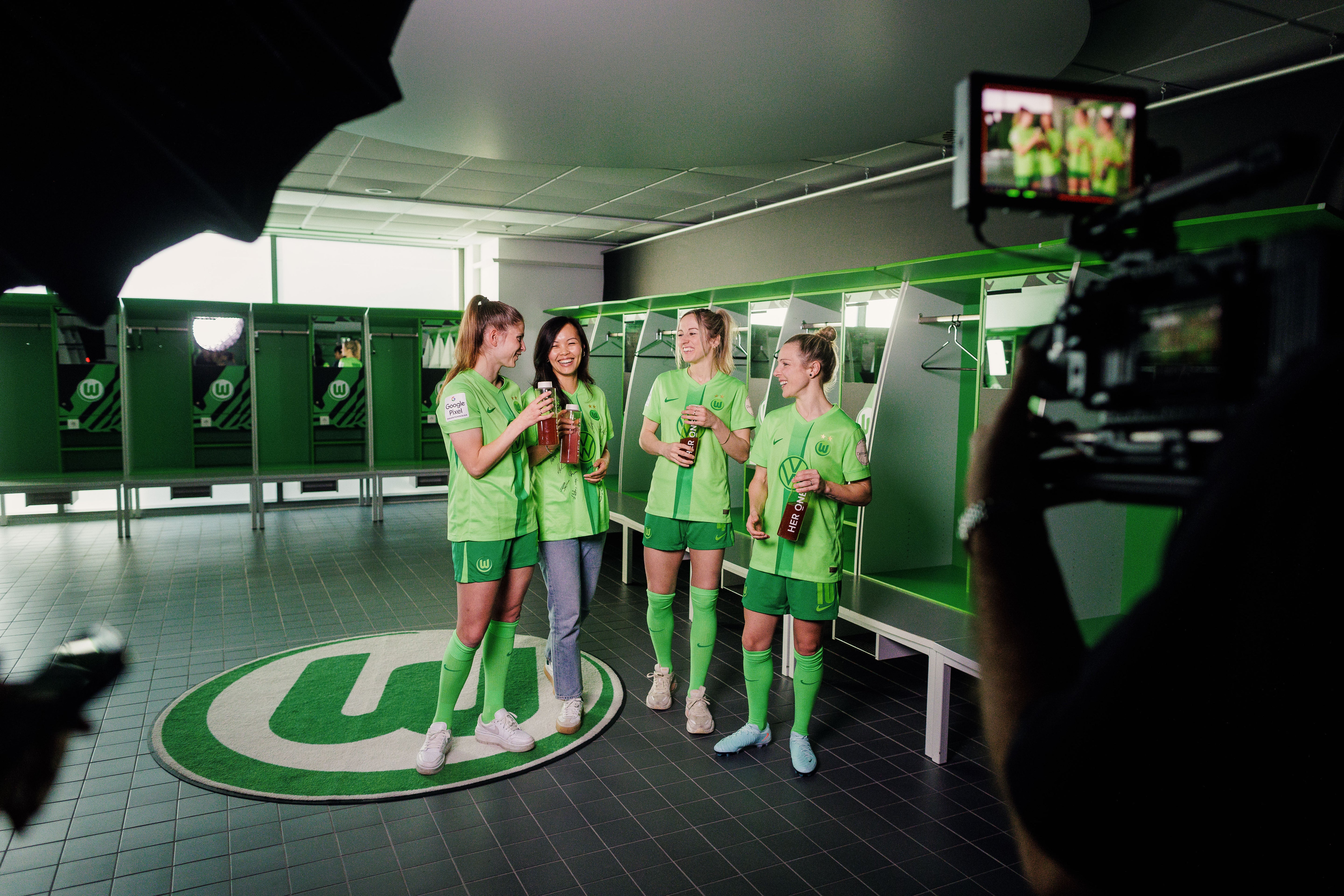 Four women in green jerseys stand in a changing room, smiling and posing with bottles in their hands.