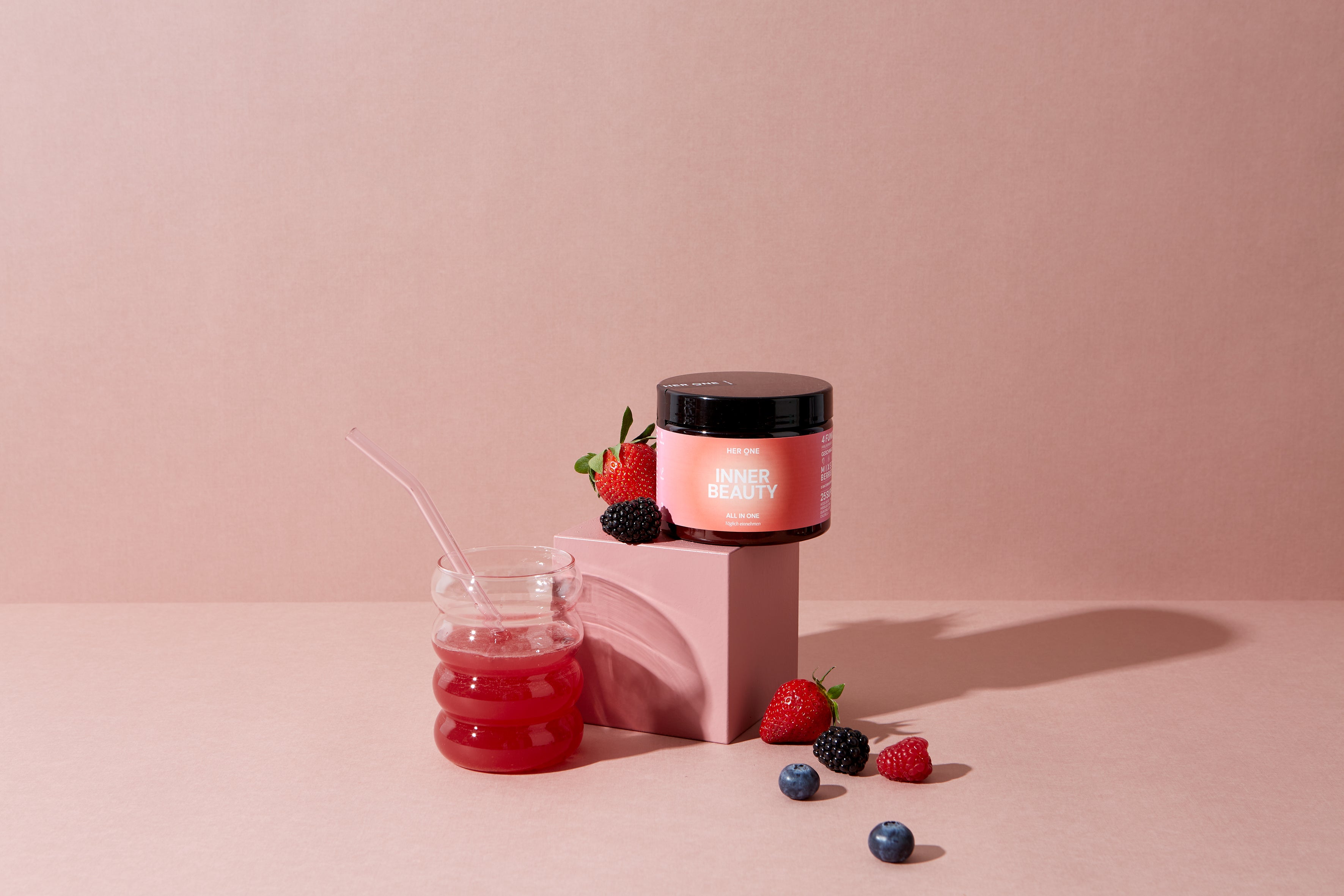 A glass with a red drink, a pink product container and various berries on a pink background.