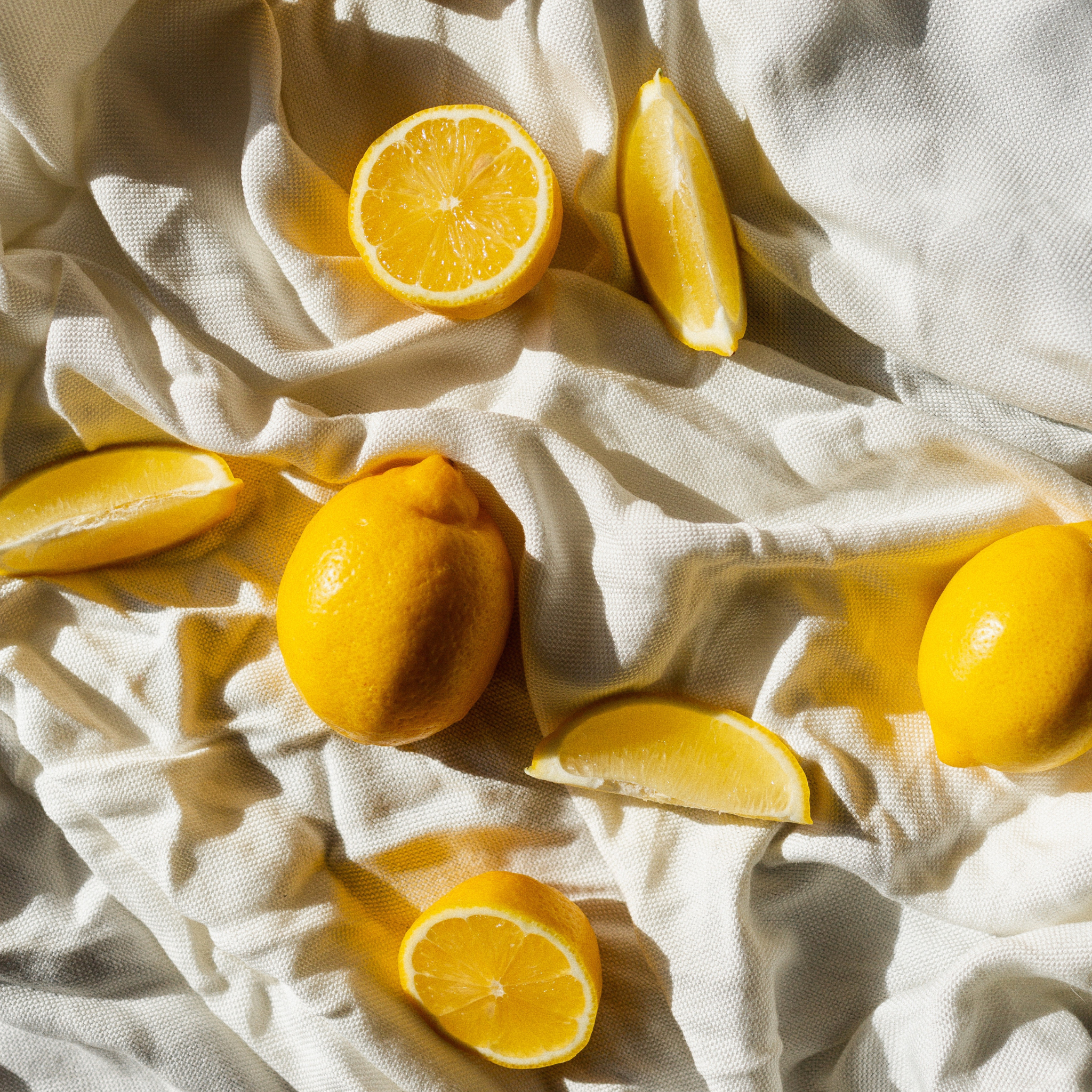 Lemons and lemon slices lie on a crumpled white cloth under sunlight.