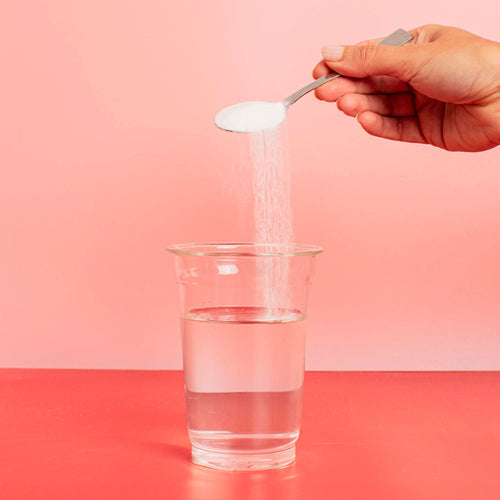 A hand holds a spoon and pours sugar into a glass of water on a red table against a pink background.