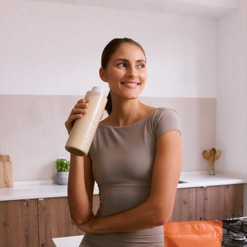 A woman stands in a modern kitchen and holds a HerOne bottle of drink in her hand, smiling and looking to the side.