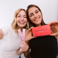 Two smiling women hold HER ONE Liquid Beauty packaging and drinking sticks in pink up to the camera.