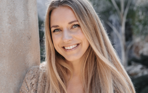 A smiling dream with long blond hair leans against a wall in the open air.