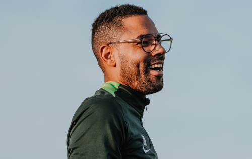 Man with glasses and beard smiling in profile, wearing a dark green jacket, against a blue sky.