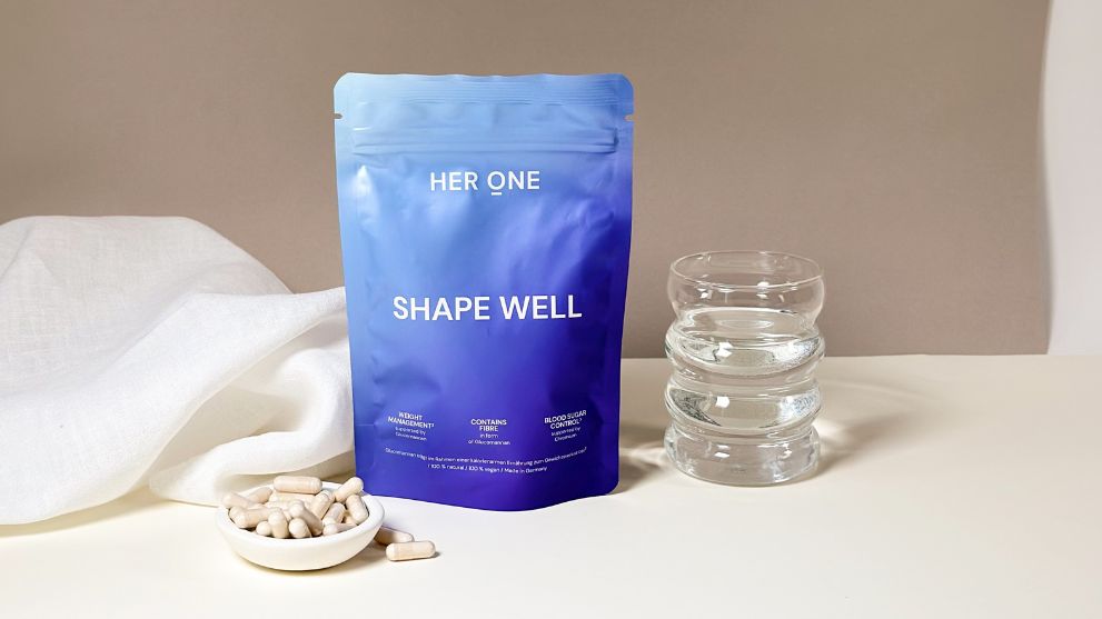 Blue-purple packaging of "Her One Shape Well" on table with white capsules in bowl and filled glass next to it.