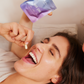 Woman lying on the bed laughing and holding a capsule while showing a bag labeled "HER ONE".