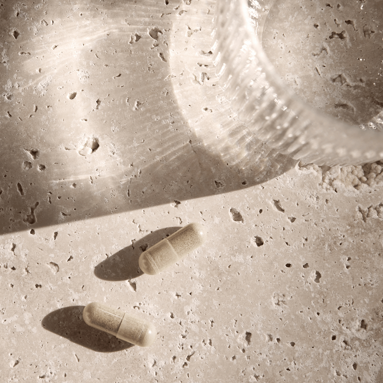 Two capsules and a glass stand on a textured surface in soft light.