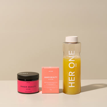Three products labeledInner Beauty" and "HER ONE" on a table, including a glass container with yellow liquid.
