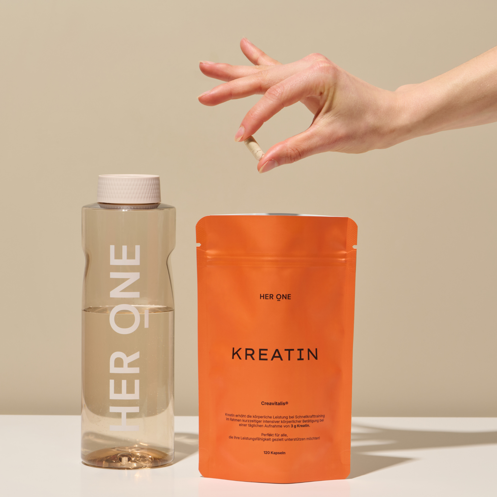 Hand holds a capsule over a packet of creatine and a bottle of water on a table.