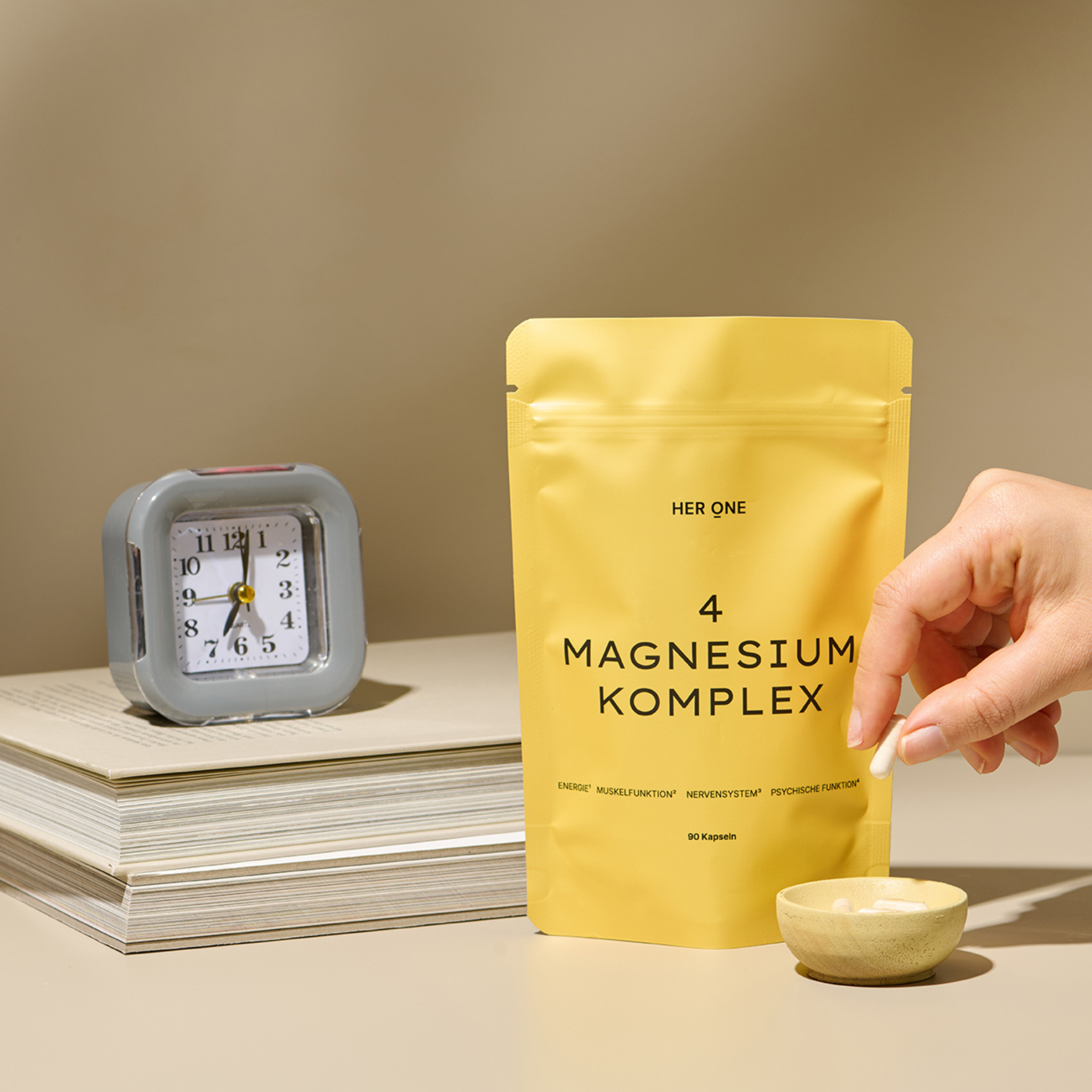 Yellow packet of "Magnesium Complex", clock on books and a hand placing a capsule in a bowl.