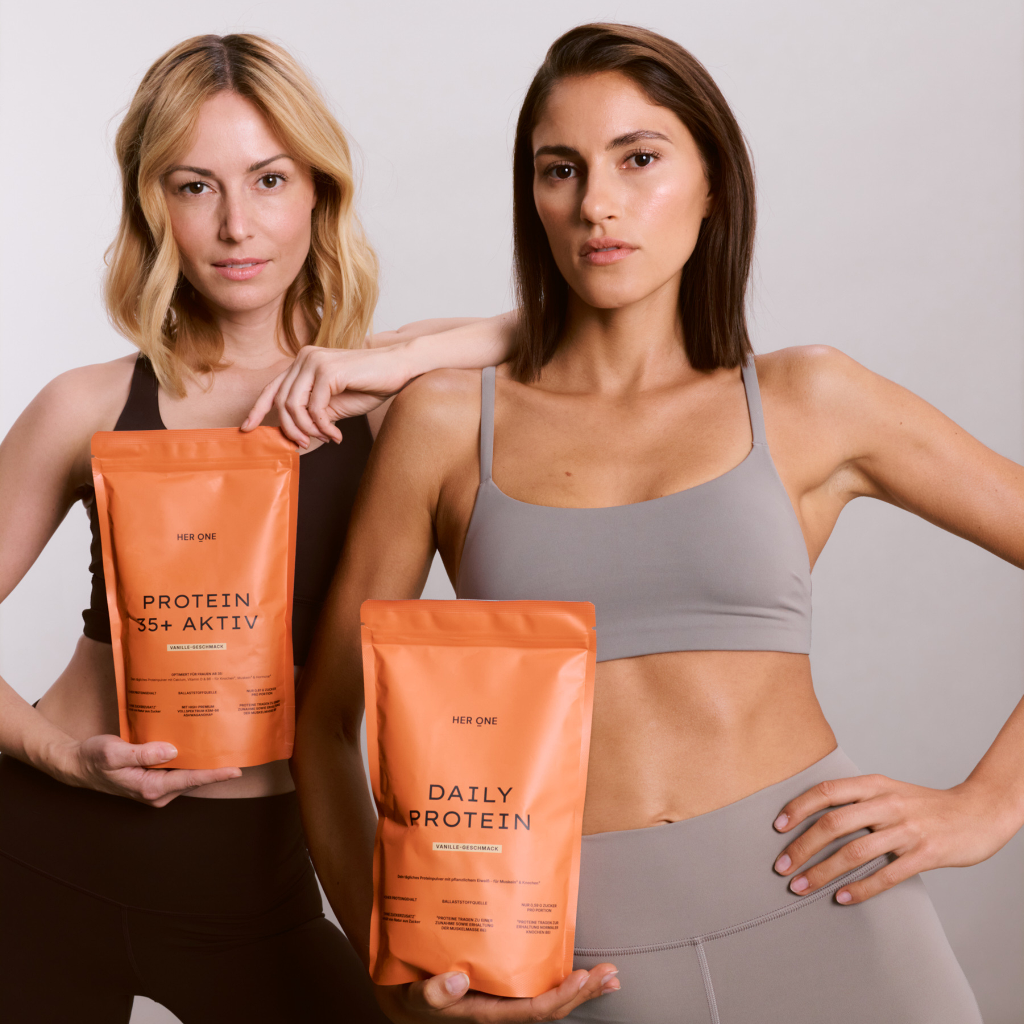 Two women in sportswear hold orange HER ONE bags labeled Daily Protein and Protein 35+ Active.