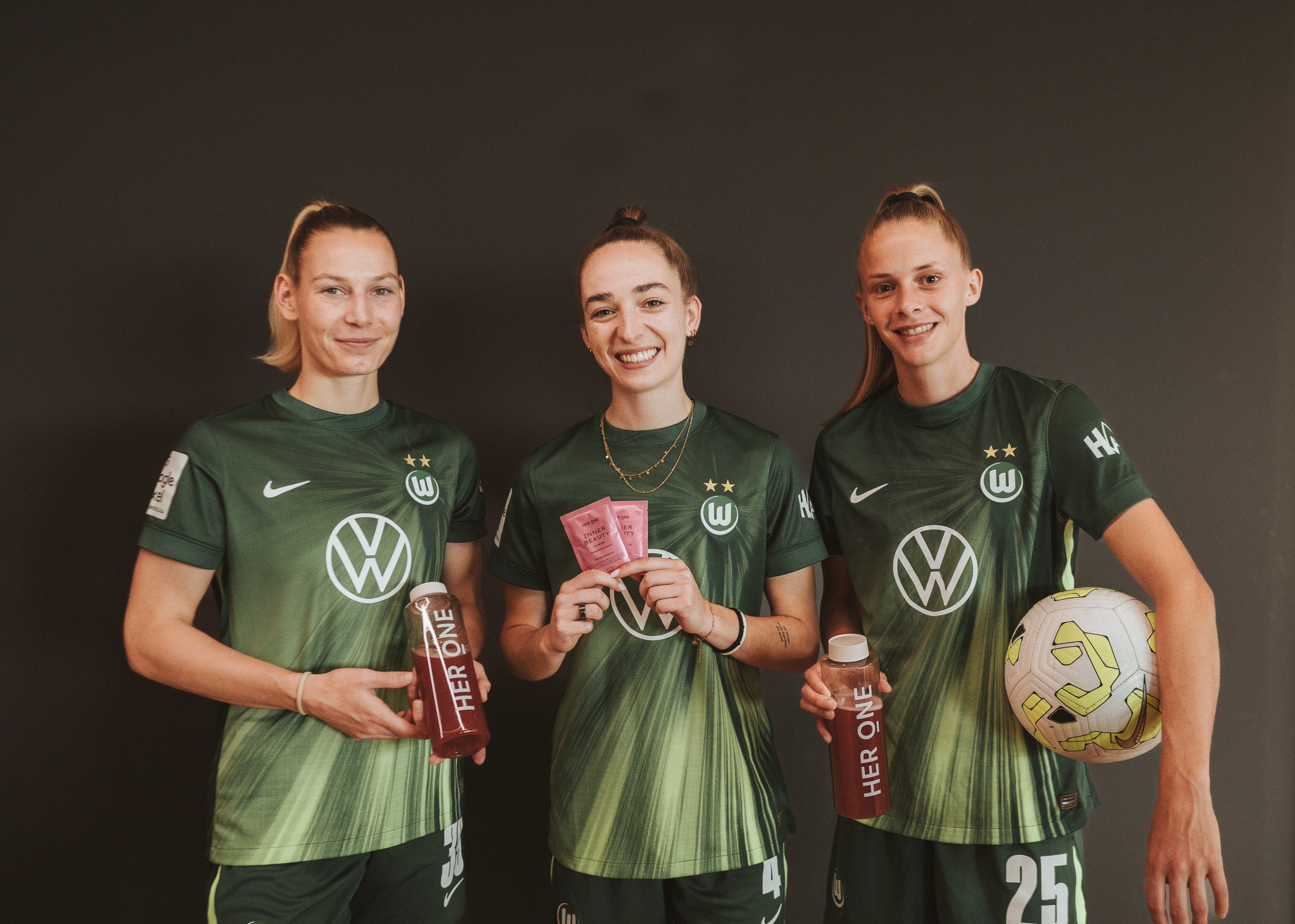 Three VfL Wolfsburg players in green jerseys hold HerOne products and smile at the camera.