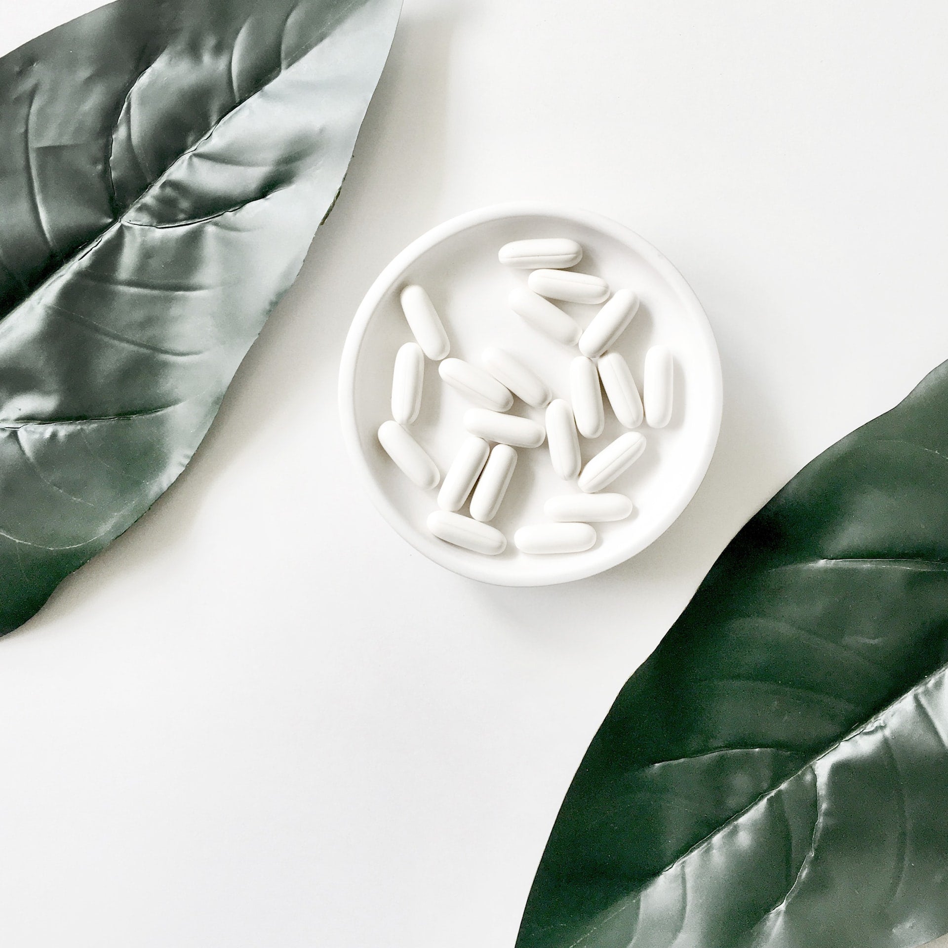 White tablets in a round bowl, surrounded by green leaves on a white background.