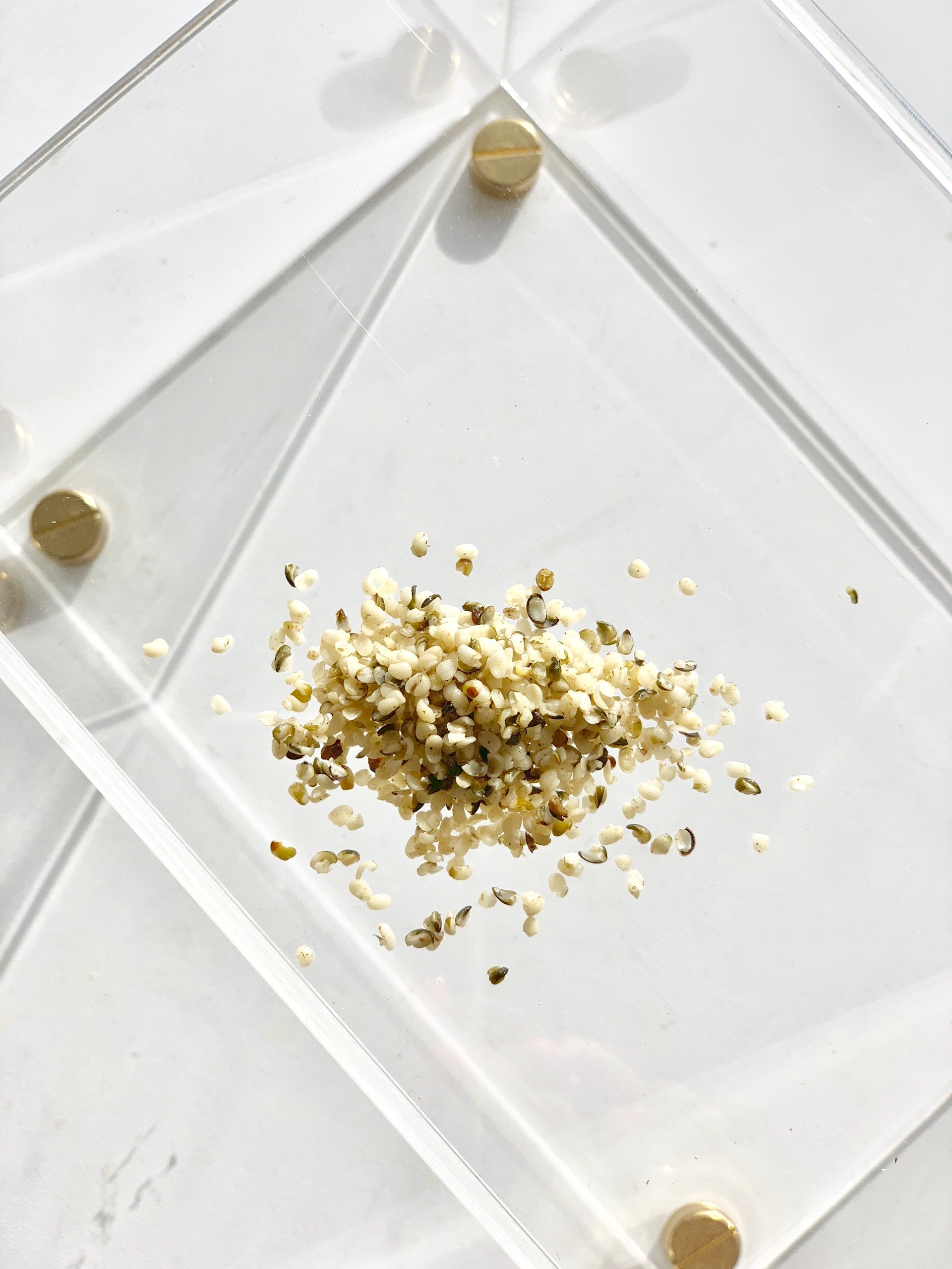 A pile of hemp seeds on a transparent, square plate with rounded corners and golden feet.