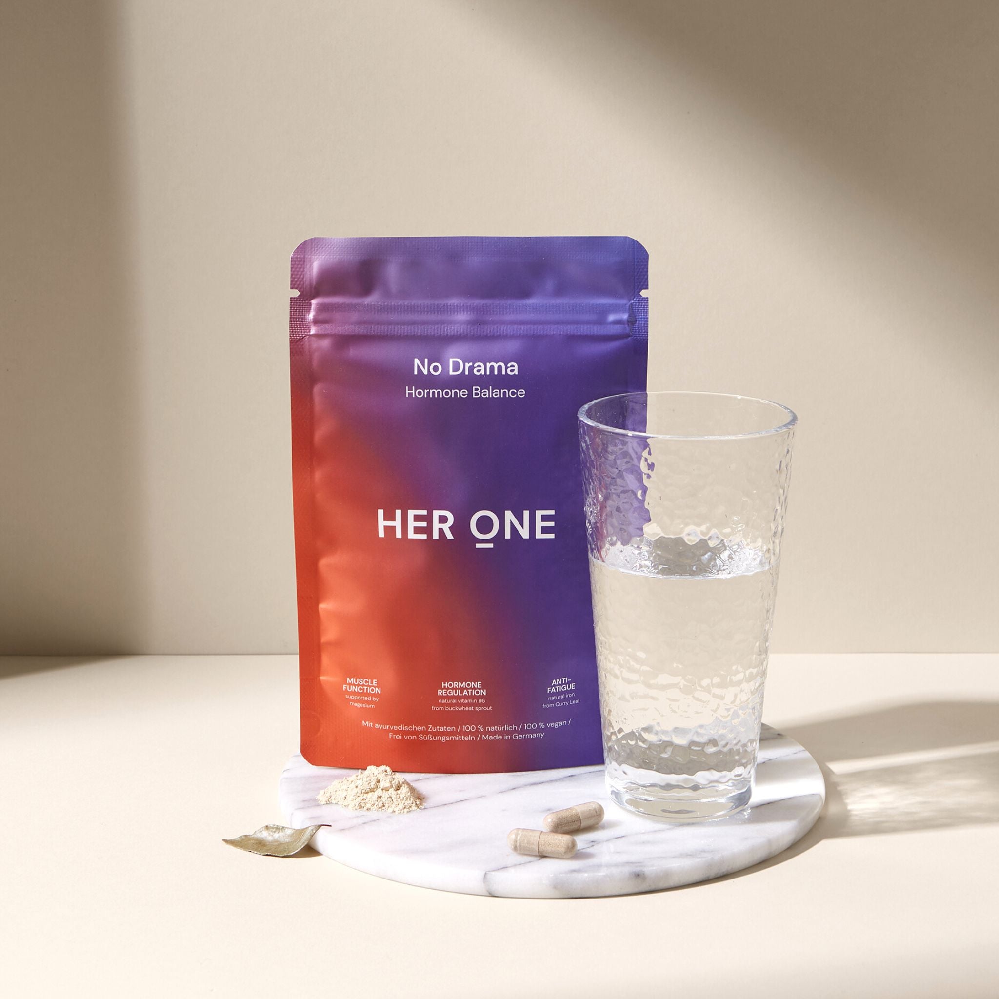A bag of "HERONE" food supplements next to a glass of water, powder and capsules on a marble tray.