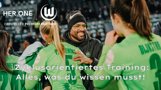 Un homme en casquette et lunettes parle à deux joueuses de football en maillot vert sur un terrain.