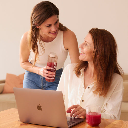 Deux femmes sourient et discutent, l'une assise devant un ordinateur portable, l'autre tenant un récipient de boissons.