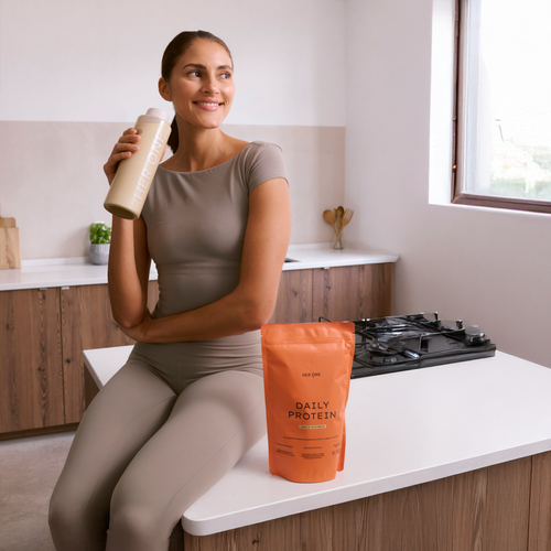 Femme en tenue de sport beige, assise dans une cuisine moderne, tenant un shake de protéines et à côté d'elle, un sachet de protéines HERONEDaily.