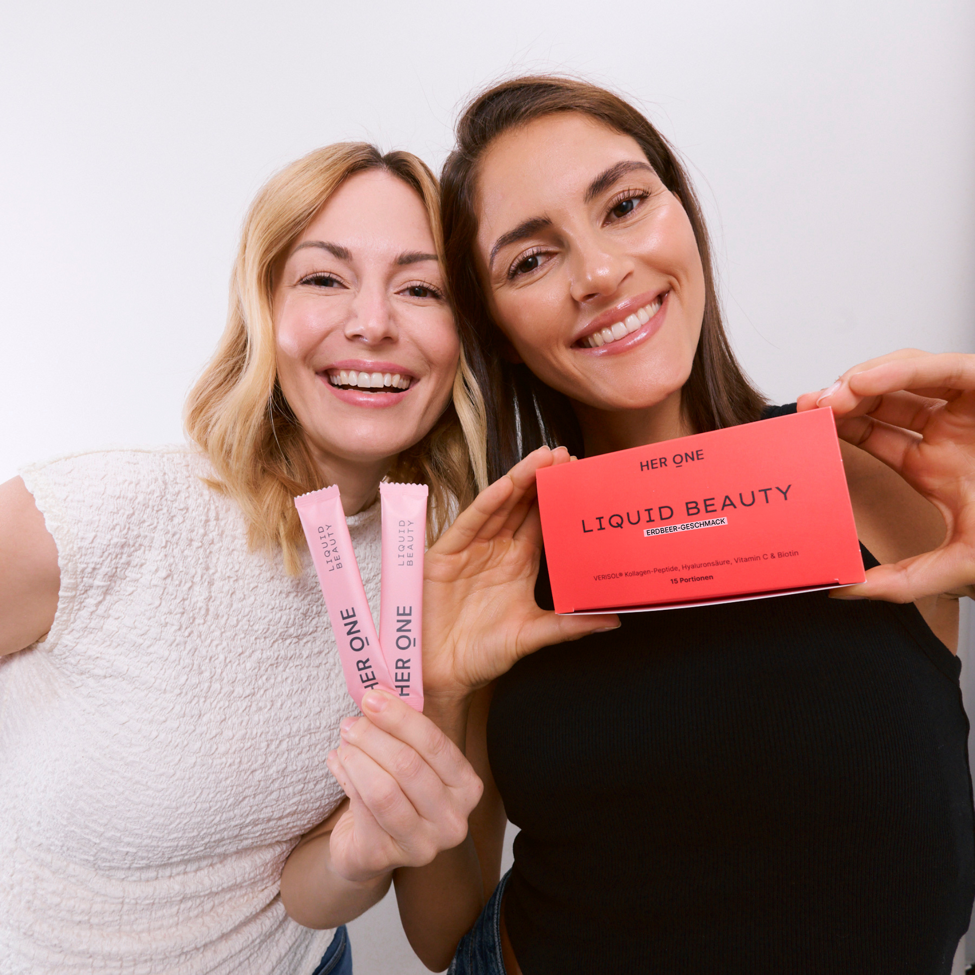 Deux femmes souriantes tiennent devant la caméra l'emballage HER ONE Liquid Beauty et des sticks à boire de couleur rose.